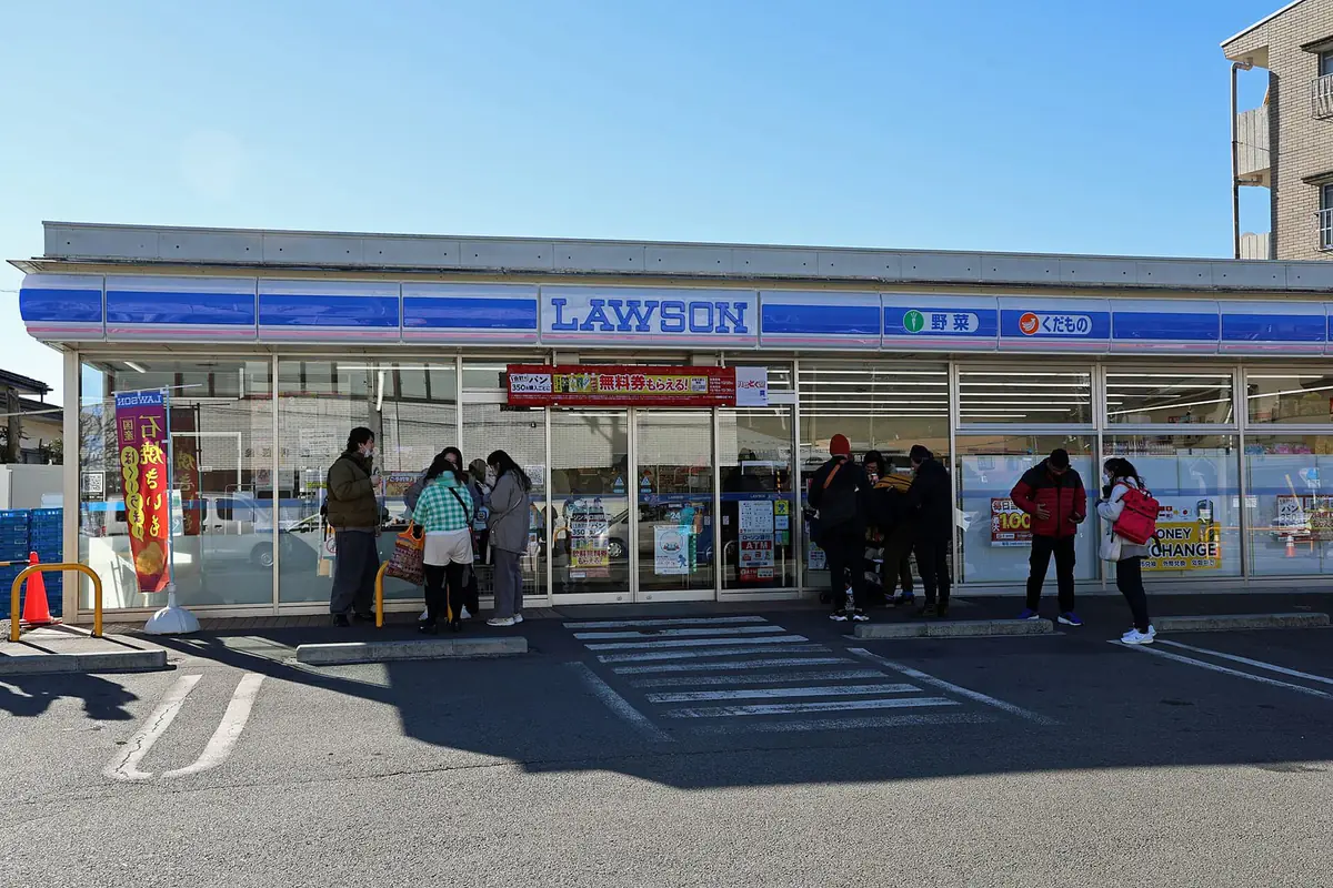 日本のローソンの店舗外観。青い看板の入口前に買い物客が立っている