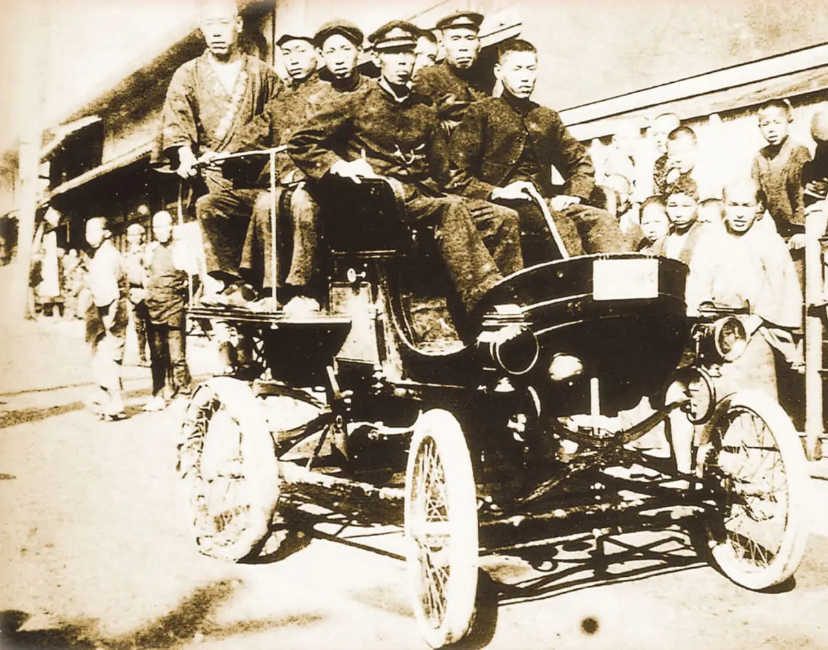 1903年に京都で運行された日本初の乗合自動車。幌のない車体の上に6人ほどが並んで座り、周りを子どもたちが囲んでいる
