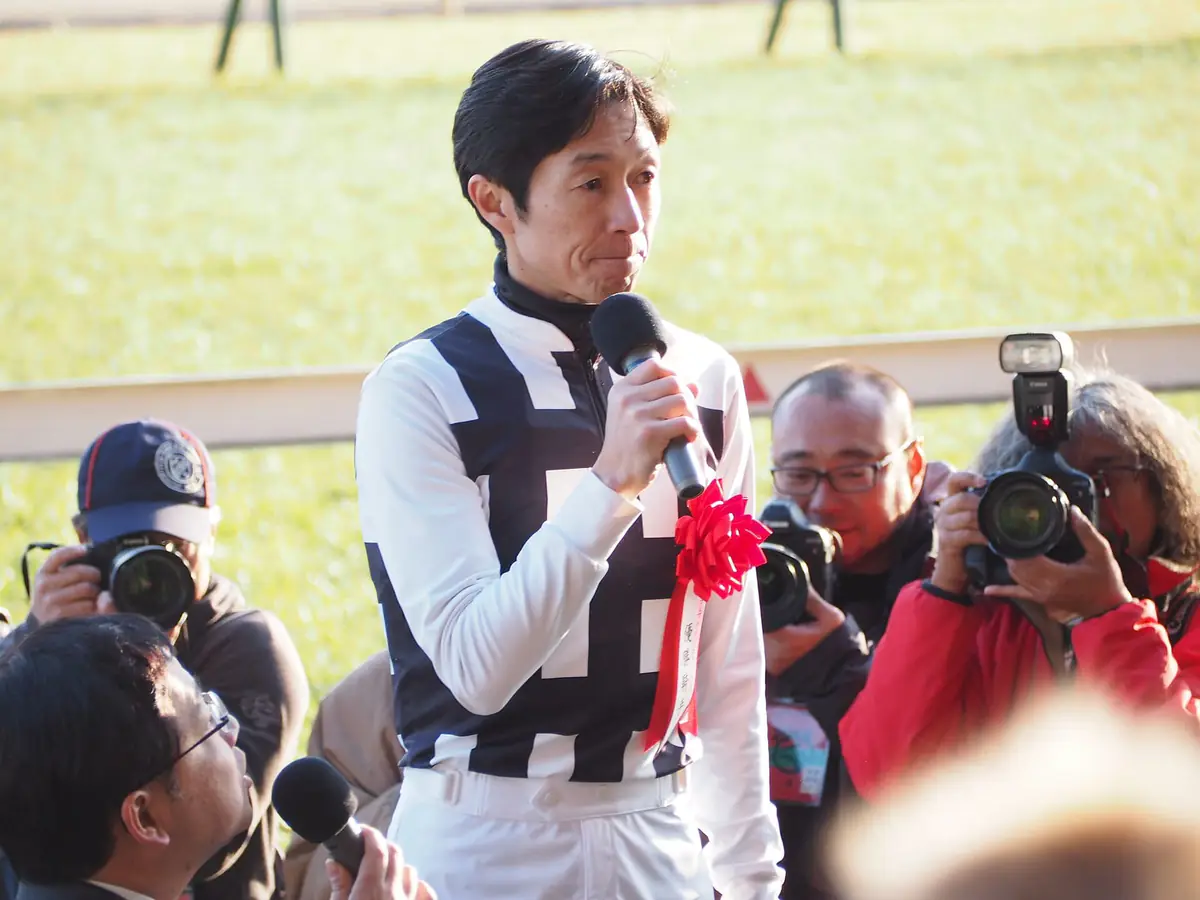 優勝騎手インタビューでマイクを持って話す武豊騎手。周りでカメラマンが撮影している